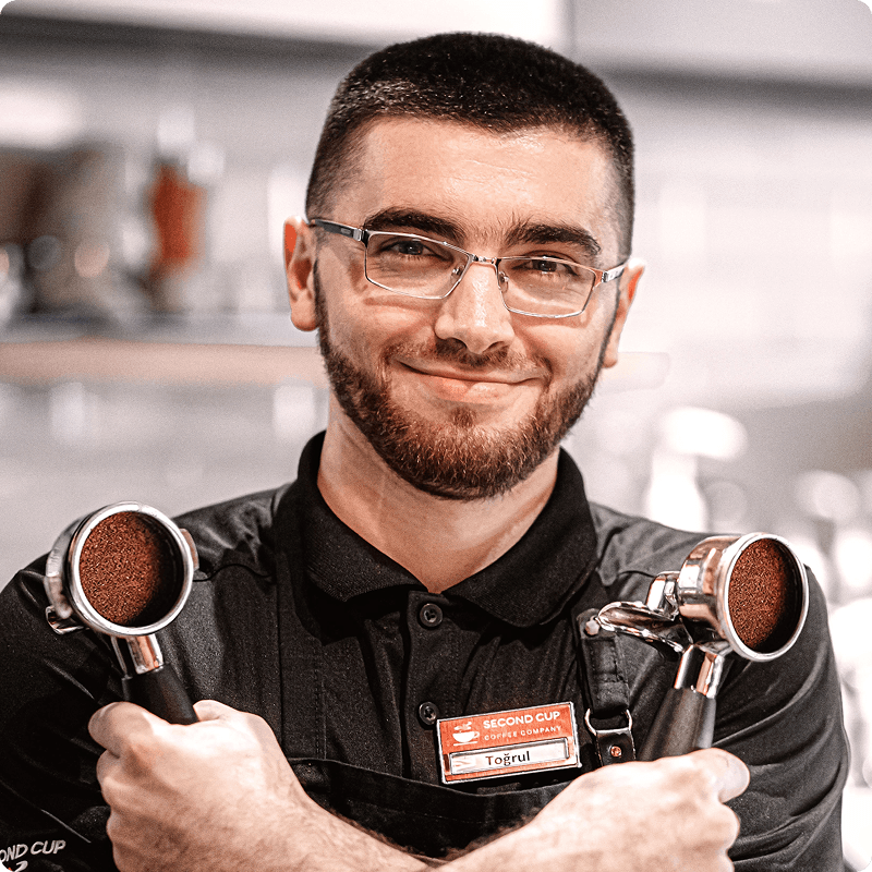 Smiling barista in glasses with a name badge, crossing his arms over two portafilters