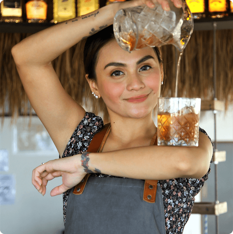 Bartender in a patterned top pouring a drink over her shoulder into a glass
